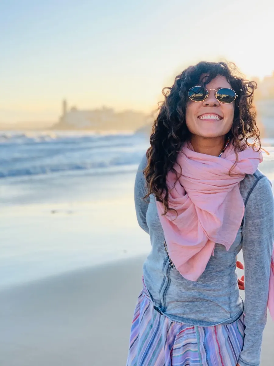 Eli Lozano laughing on Sitges beach at golden hour, Sant Bartomeu behind her
